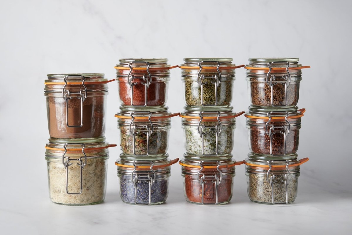 An overhead, top-down view of various glass jars and wooden bowls containing colorful salts and dried herbs on a dark wooden surface. Scattered loose botanicals and peppercorns create a rustic, textured culinary scene.