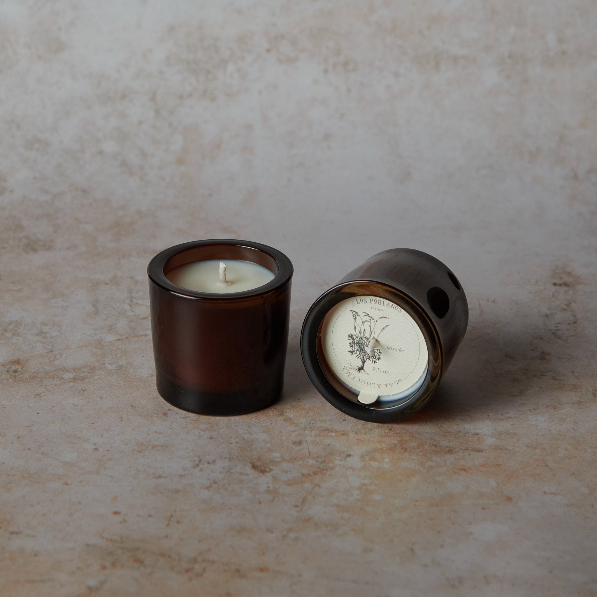 Two brown glass candle holders with candles on a textured beige surface