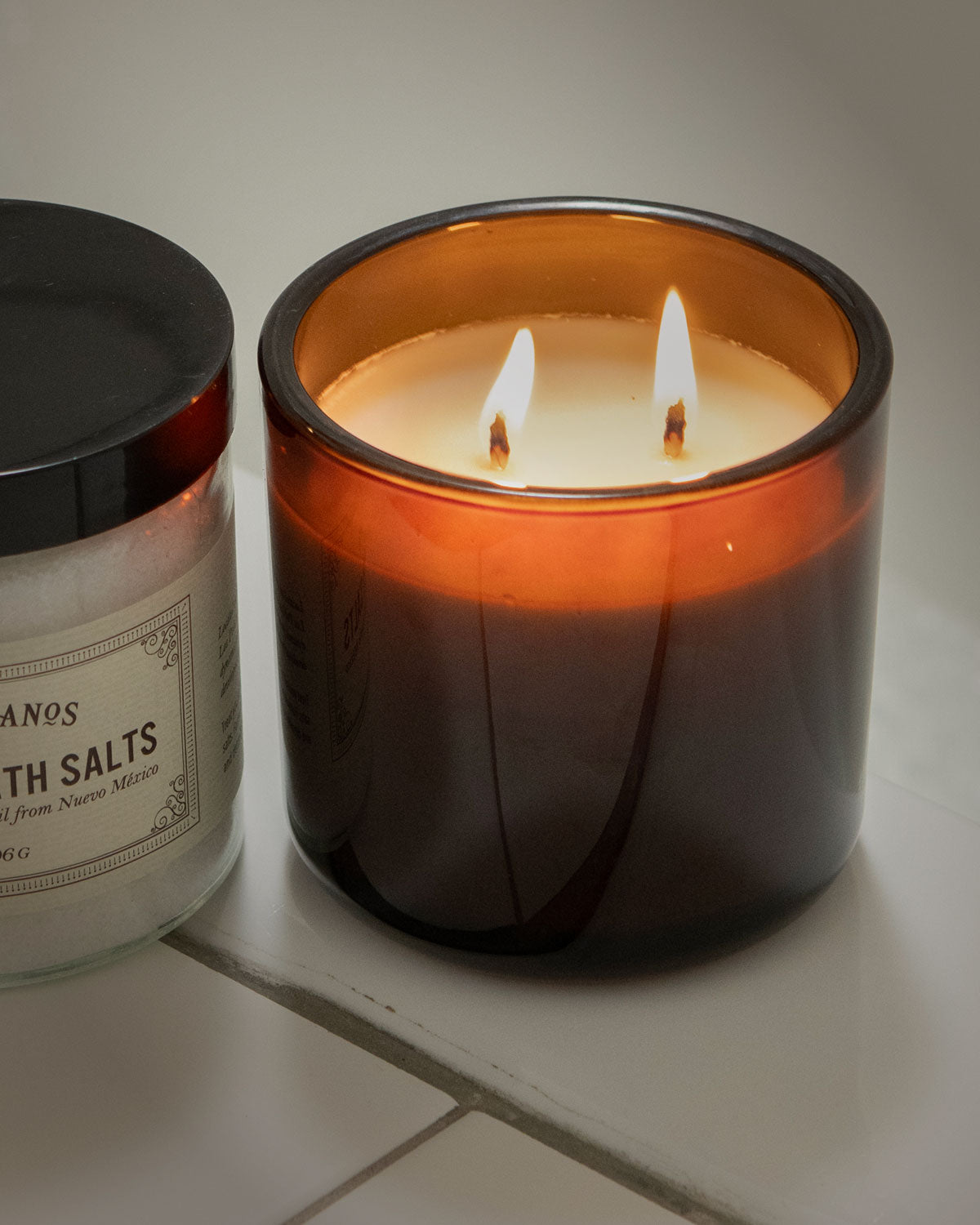 Candle with a lit wick next to a jar labeled 'Bath Salts' on a light surface.