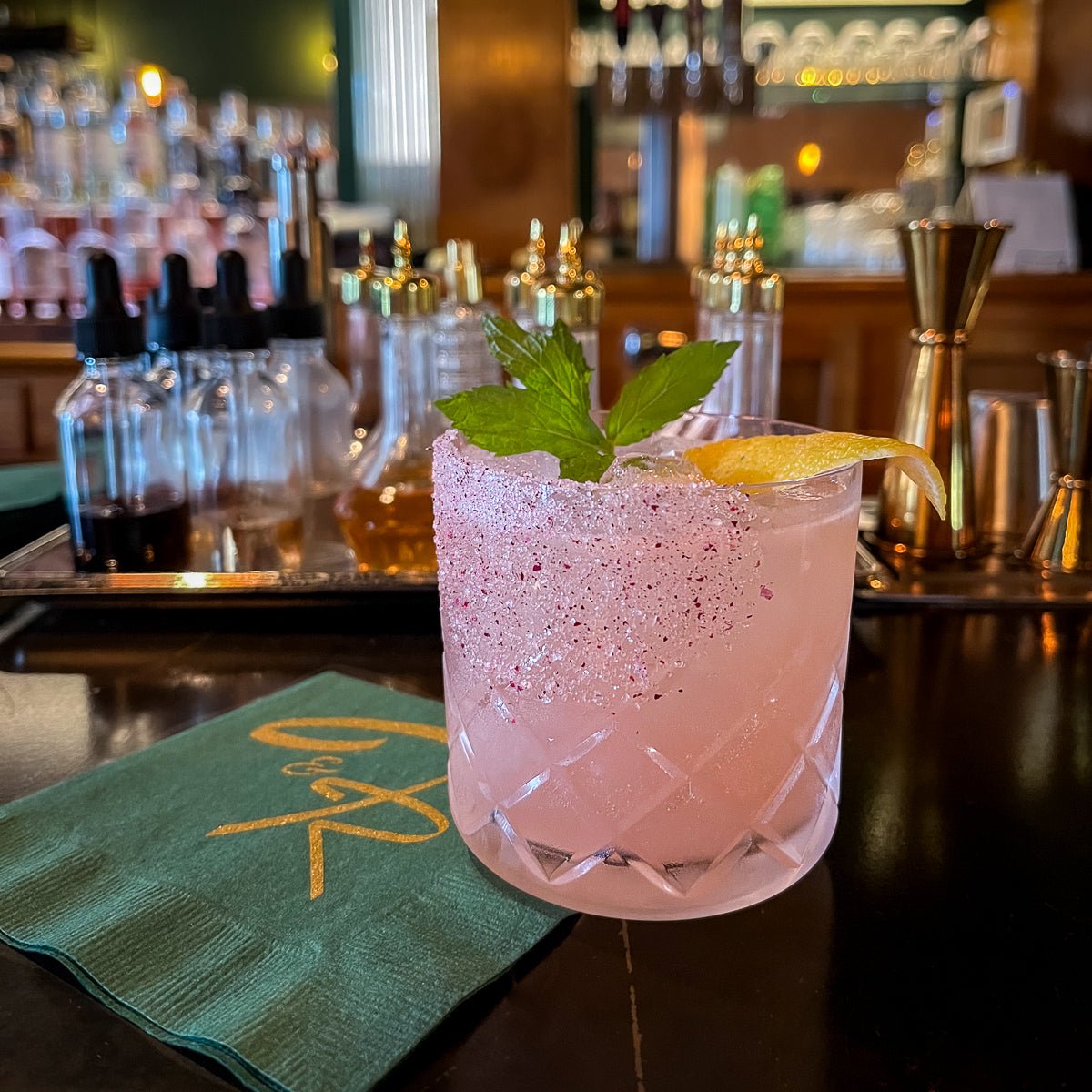 Pink cocktail with a lemon twist and mint leaf on a bar counter with bottles and tools in the background.