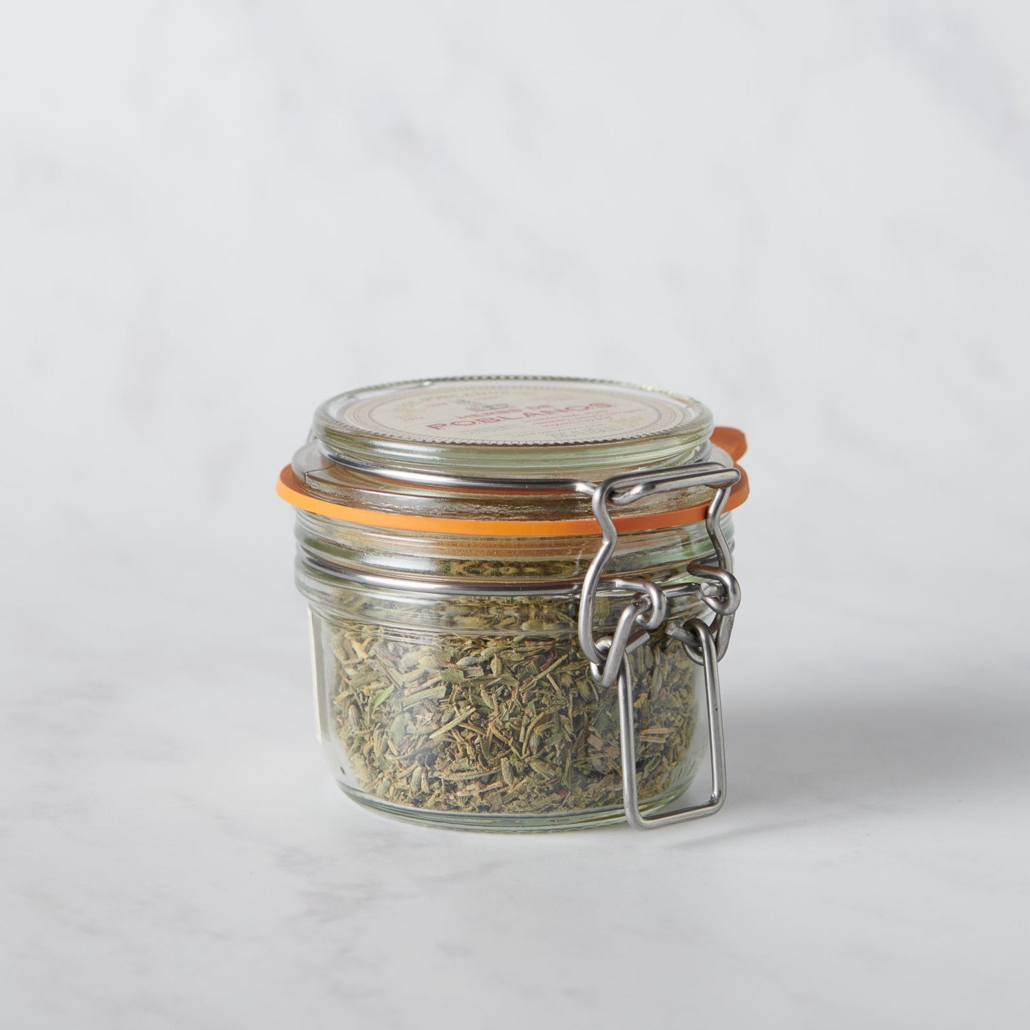 Small glass jar with a metal clasp full of French Herbs on a white background