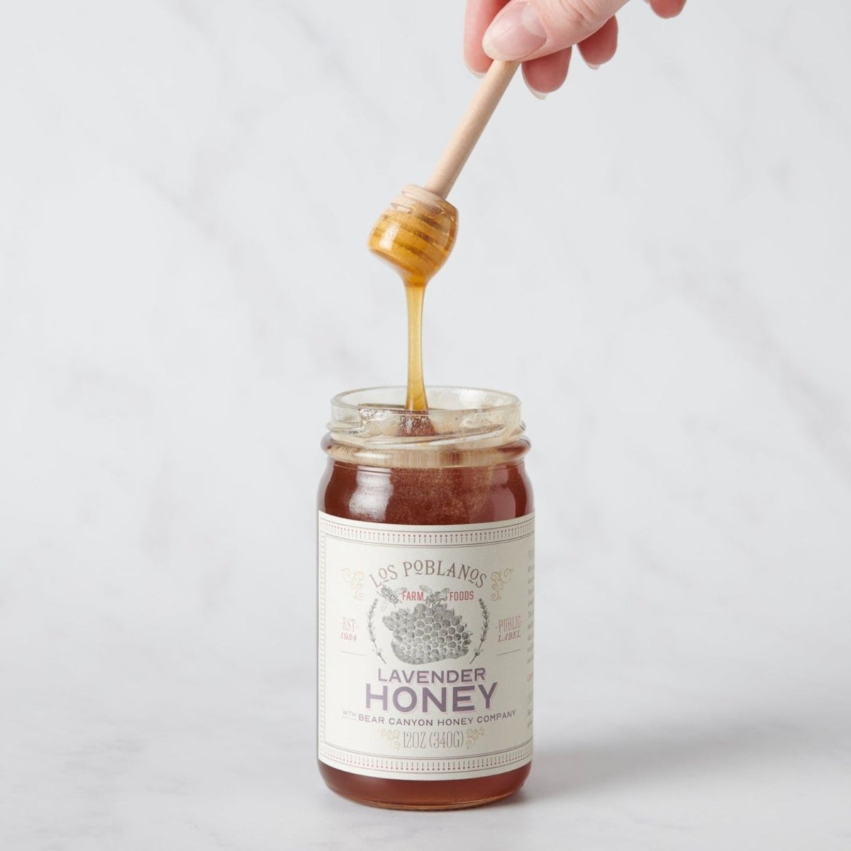 A jar of lavender honey with a hand holding a honey dipper above it, showing the product's texture and color | Lavender Honey | Los Poblanos Farm Shop