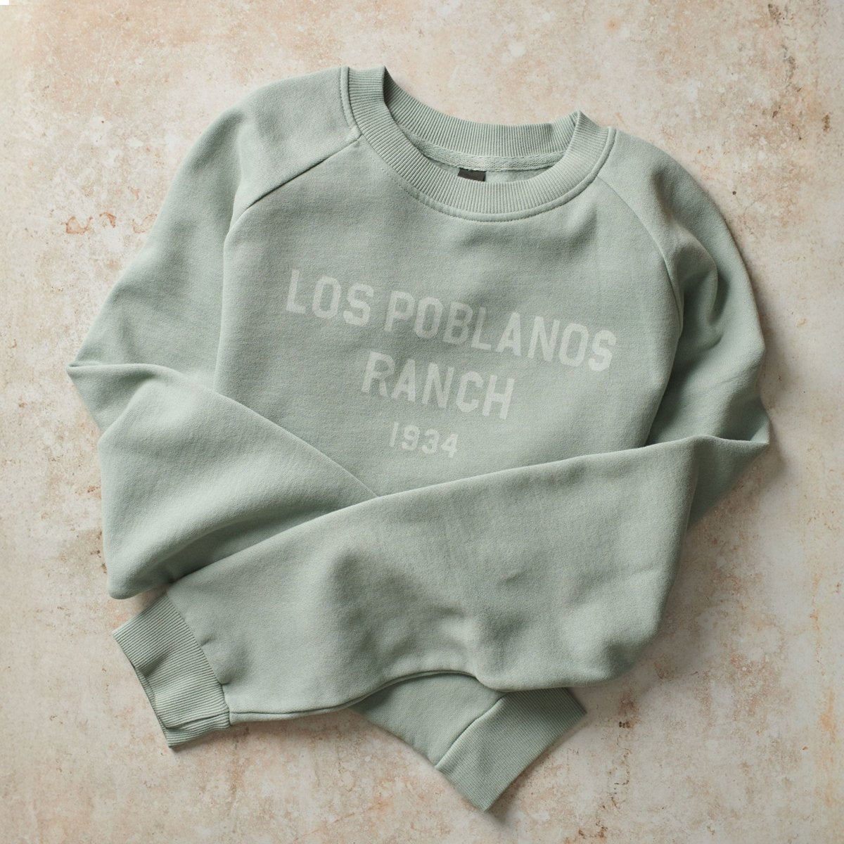 Green sweatshirt with 'Los Poelanos Ranch 1934' text on a beige surface