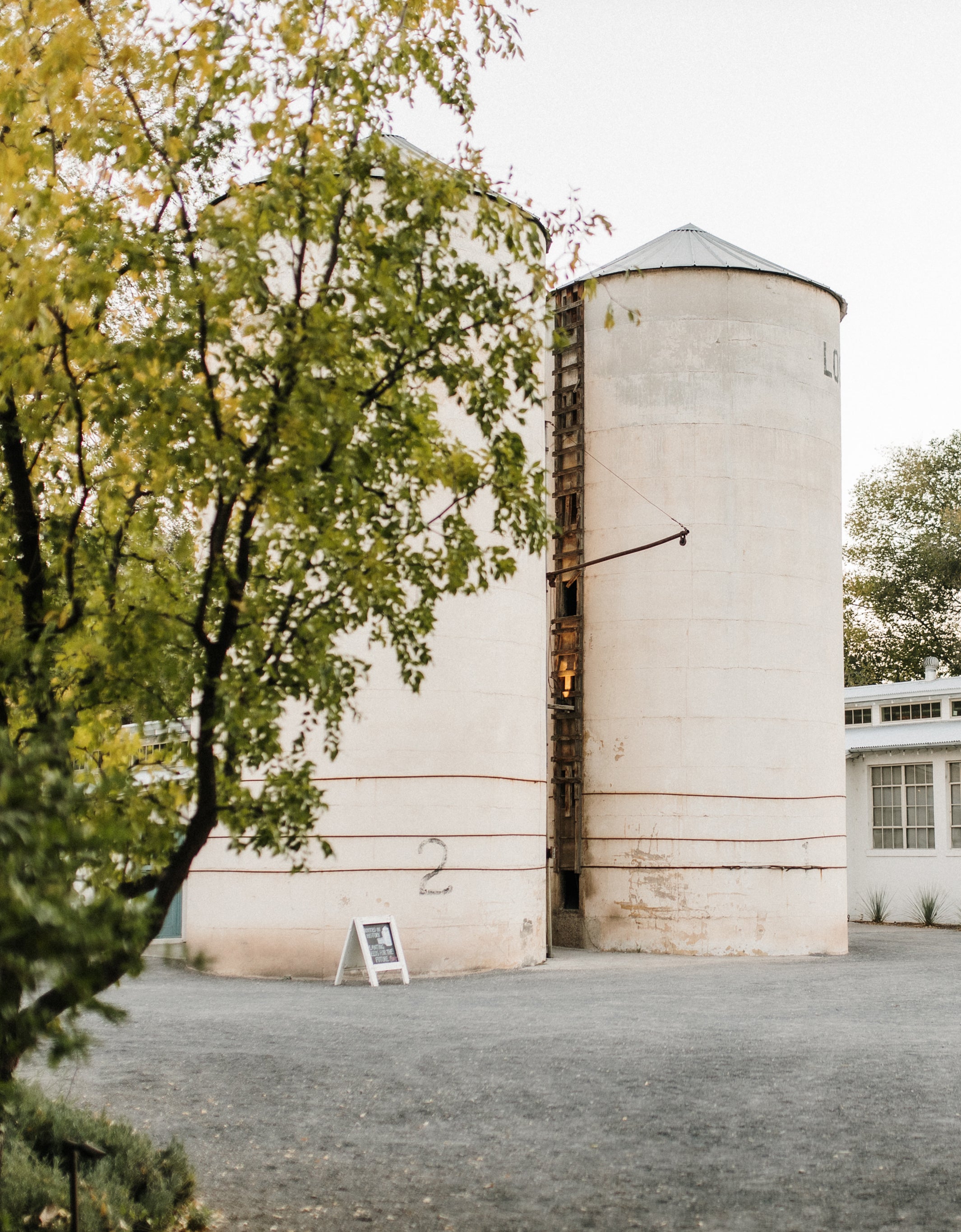 Large white silo with a smaller cylindrical structure, surrounded by trees and a building.