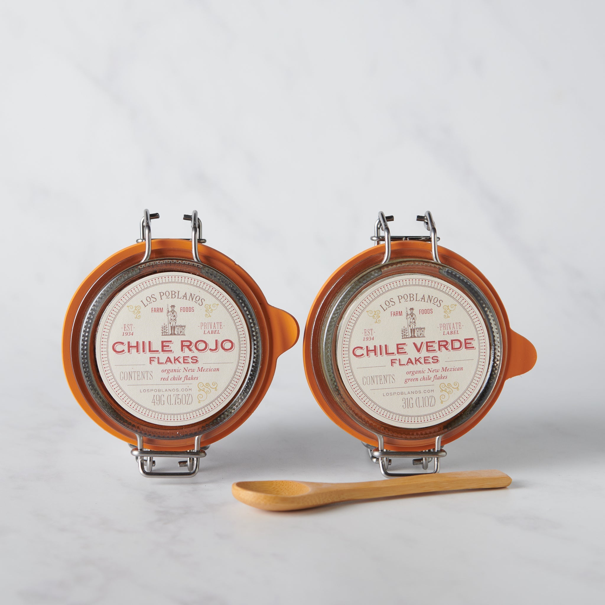 Two round containers labeled 'Chile Rojo Flakes' and 'Chile Verde Flakes' with a wooden spoon on a light gray background.
