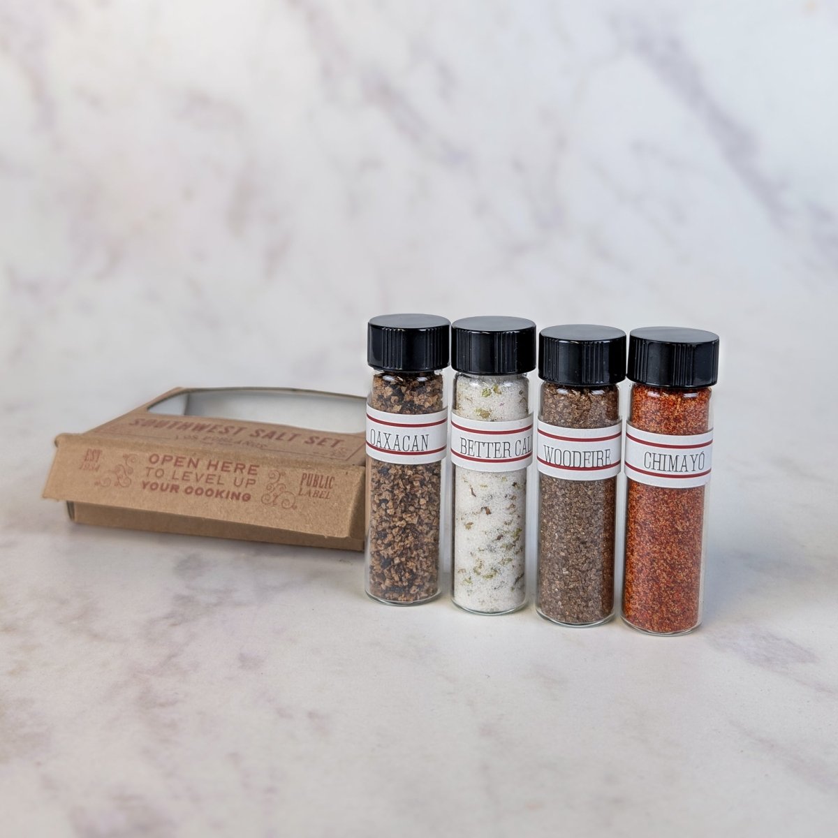 A seasoning salt set with four clear glass bottles containing black, white, brown, and red spices, standing next to a brown cardboard box on a white marble surface.