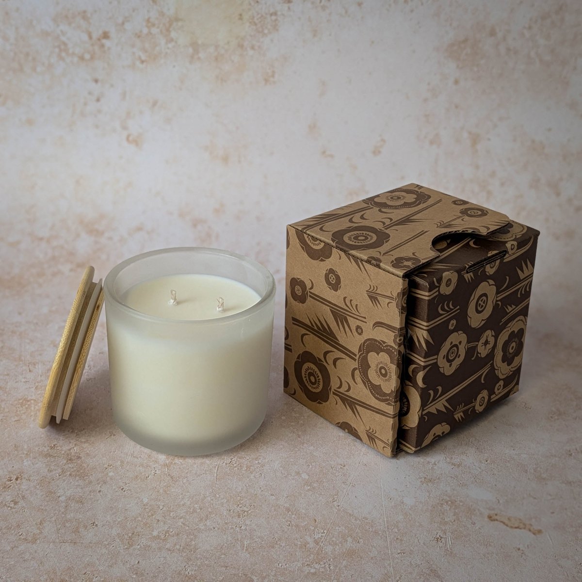 Glass candle with wooden lid next to a patterned cardboard box on a beige surface.