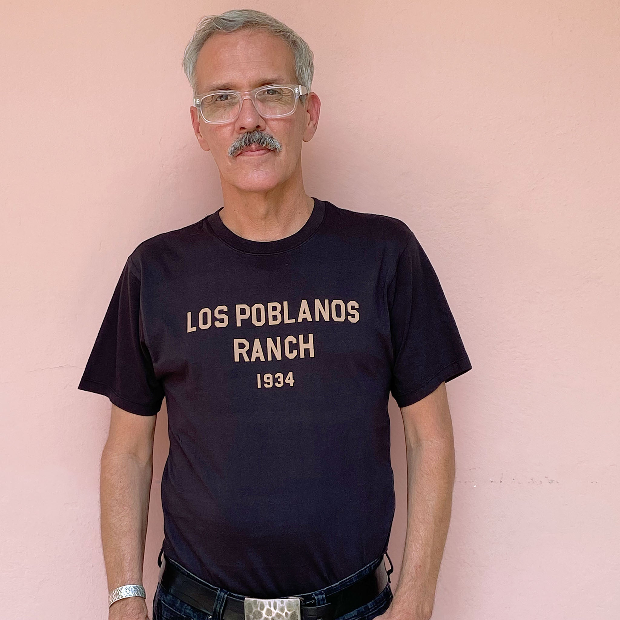 Man wearing a black t-shirt with 'Los Poblanos Ranch 1934' text against a pink background