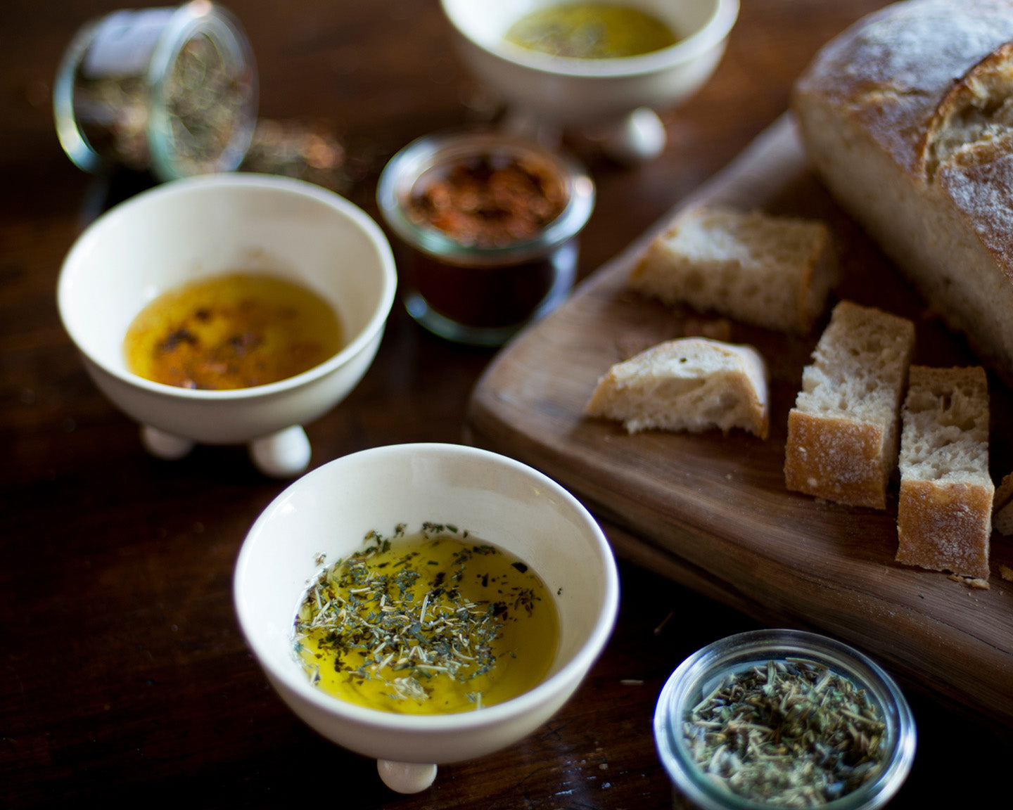 Bread slices on a wooden board with bowls of olive oil and herbs on a wooden surface.
