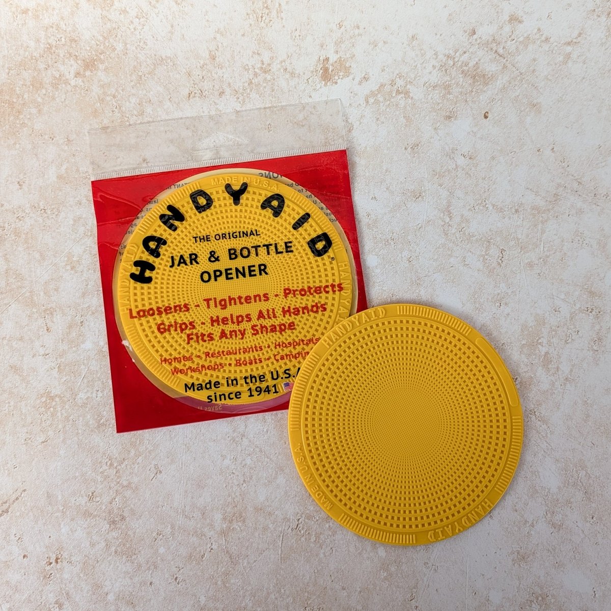 Yellow Handy Aid jar opener on a beige surface with red packaging in the background.
