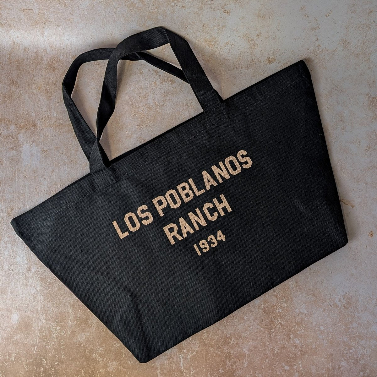 Black tote bag with 'Los Poblanos Ranch 1934' text on a beige surface
