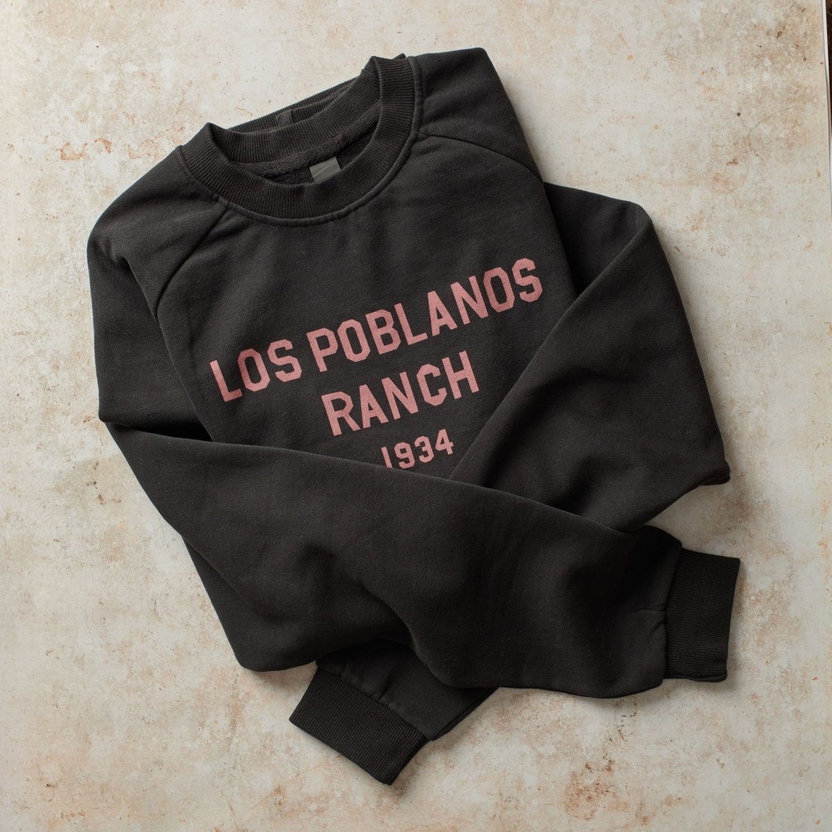 Black sweatshirt with 'Los Poblanos Ranch 1934' text on a beige surface
