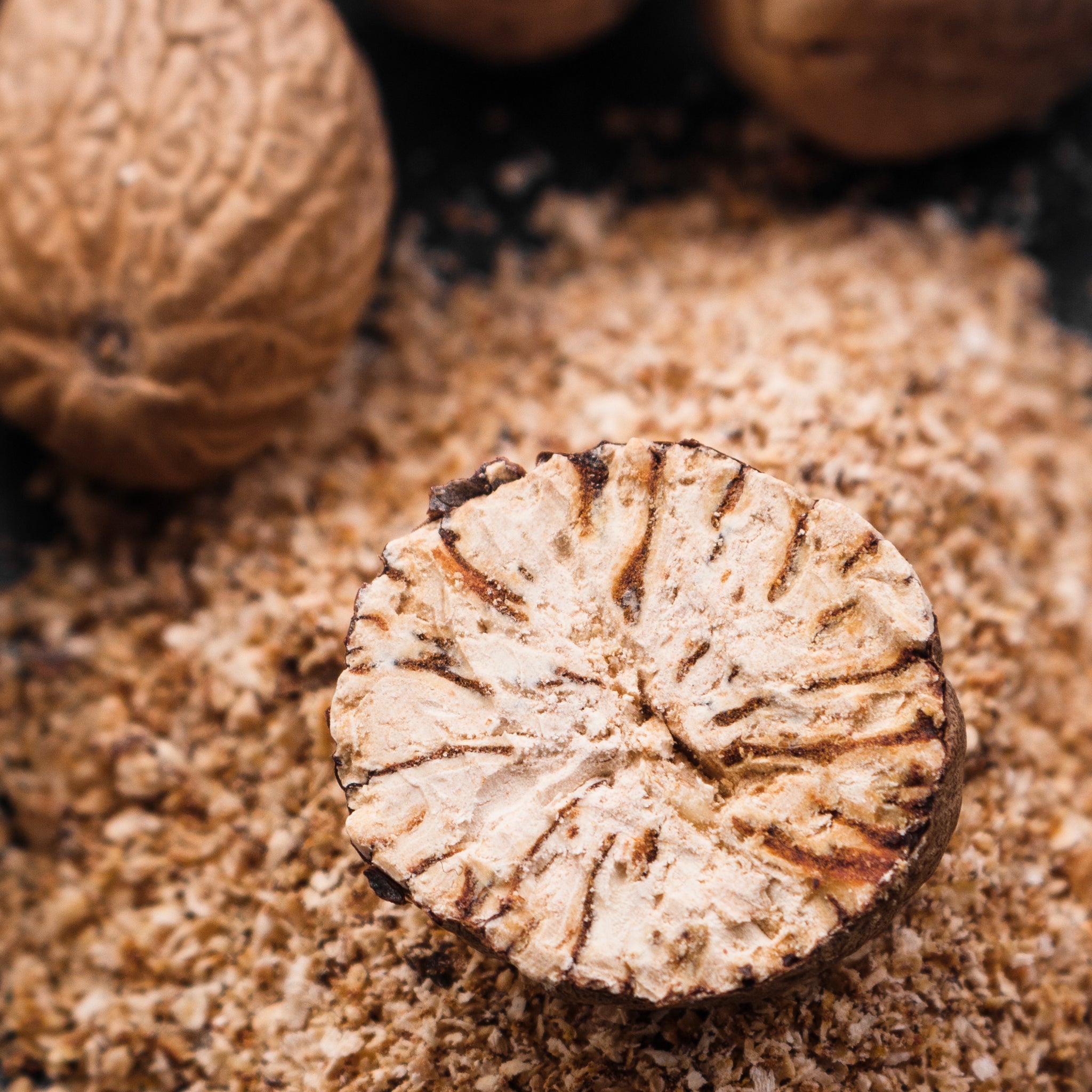 Close-up of nutmeg cut in half on a textured surface with whole nutmeg in the background