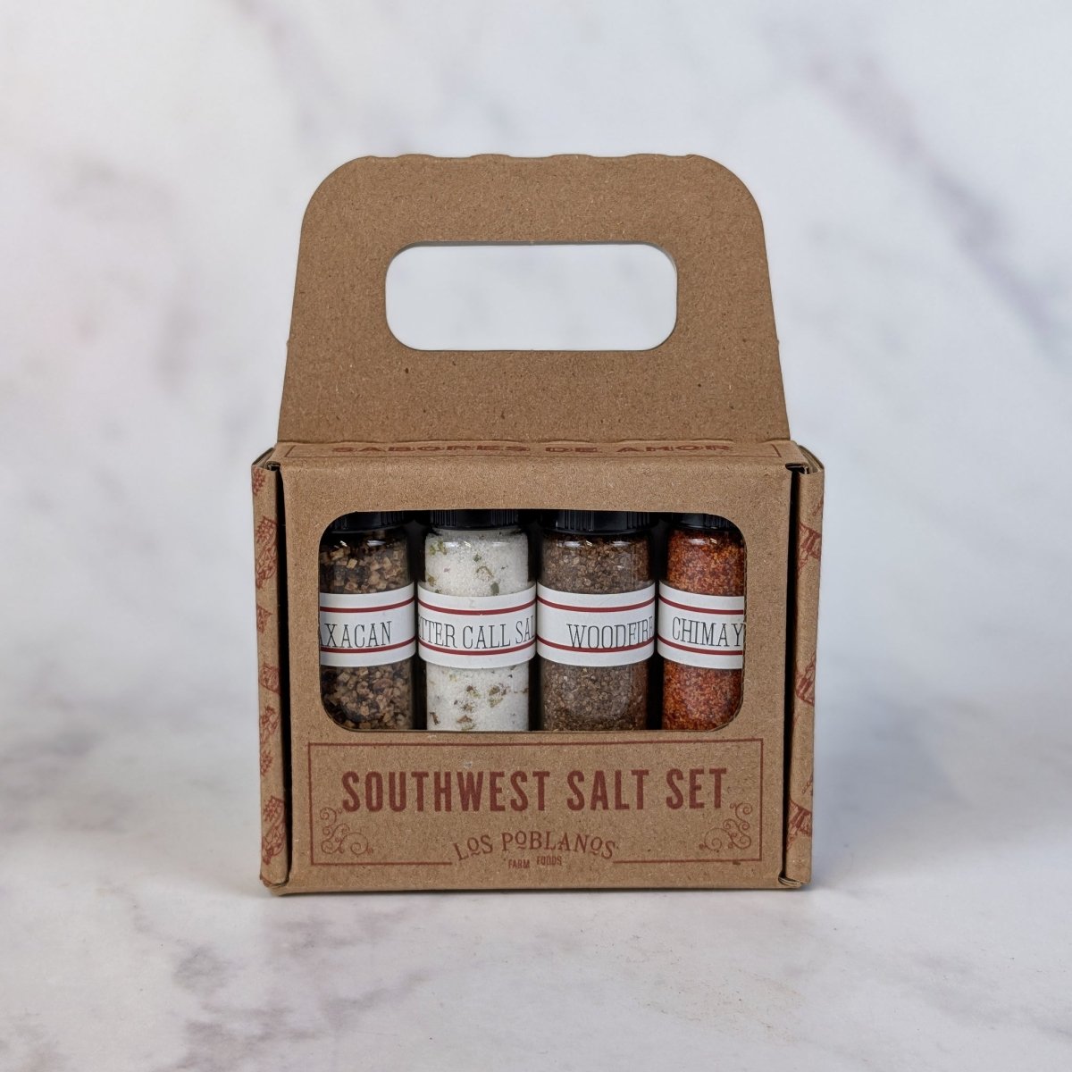 A Southwest Salt Set in a brown kraft paper carry box with a handle, containing four small glass spice jars labeled Woodfire, Oaxacan, Better Call Sal and Chimayo, on a light marble surface.