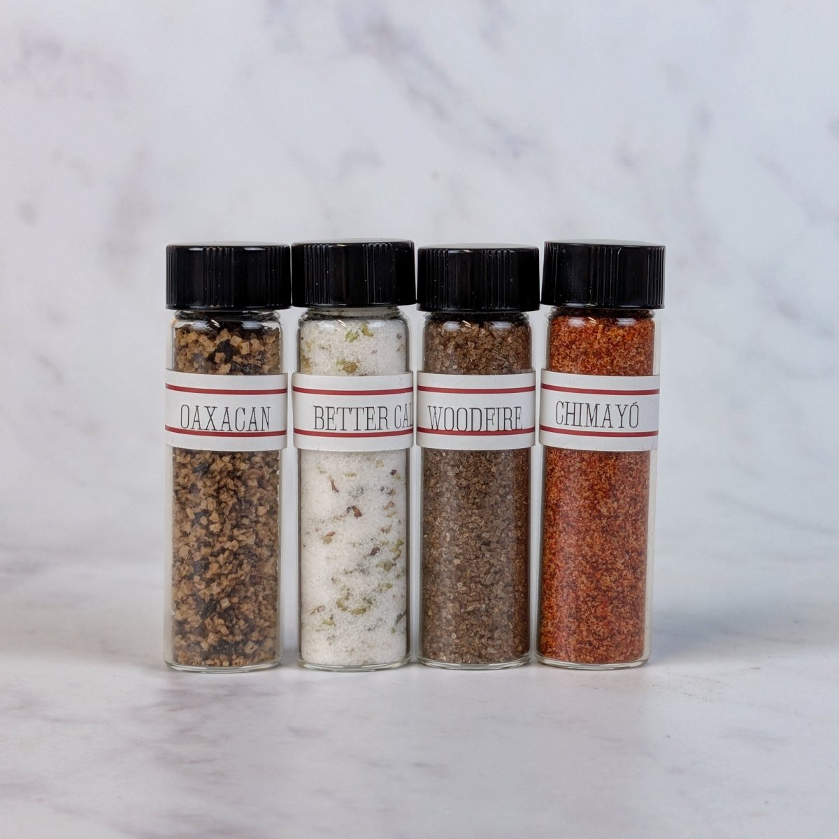 Four clear spice jars with black caps and white labels reading Woodfire, Oaxacan, Better Call Sal and Chimayo, arranged on a light marble surface against a marble background.