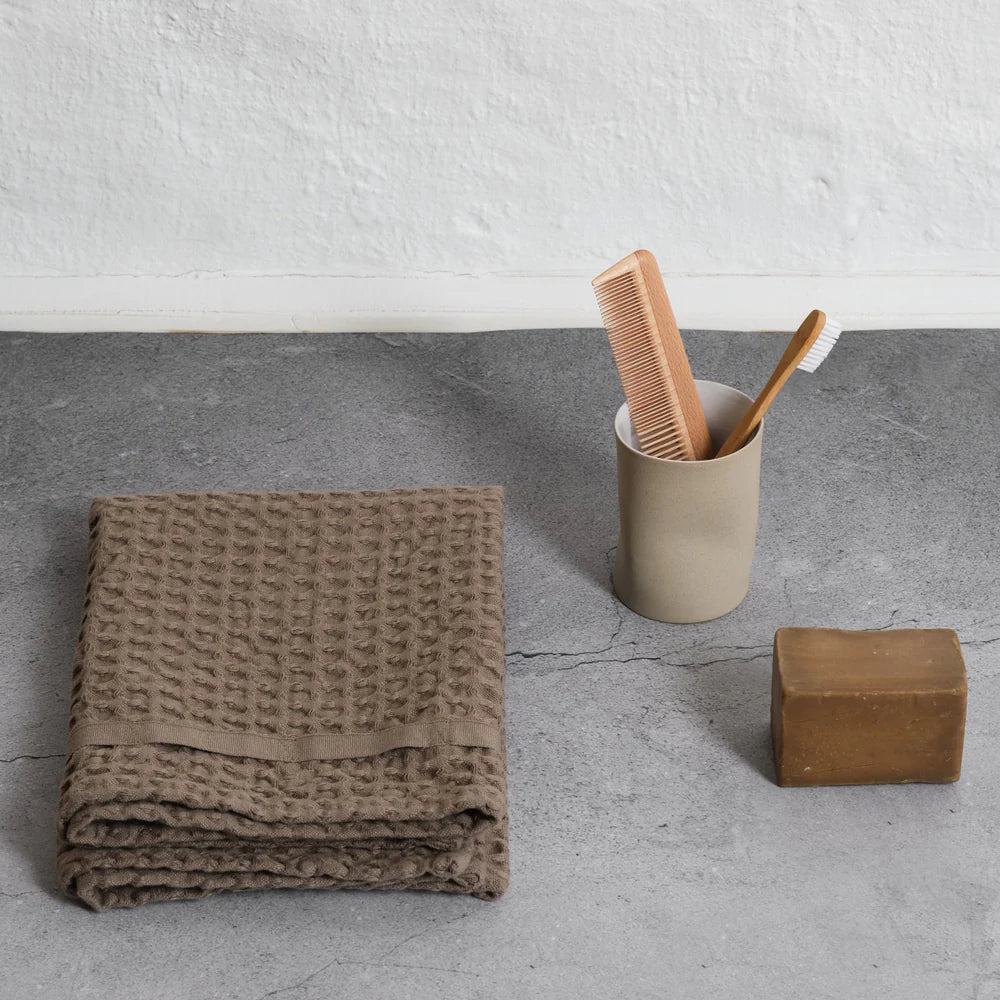 Folded brown towel, beige cup with bamboo toothbrush and comb, and brown soap bar on a gray surface.