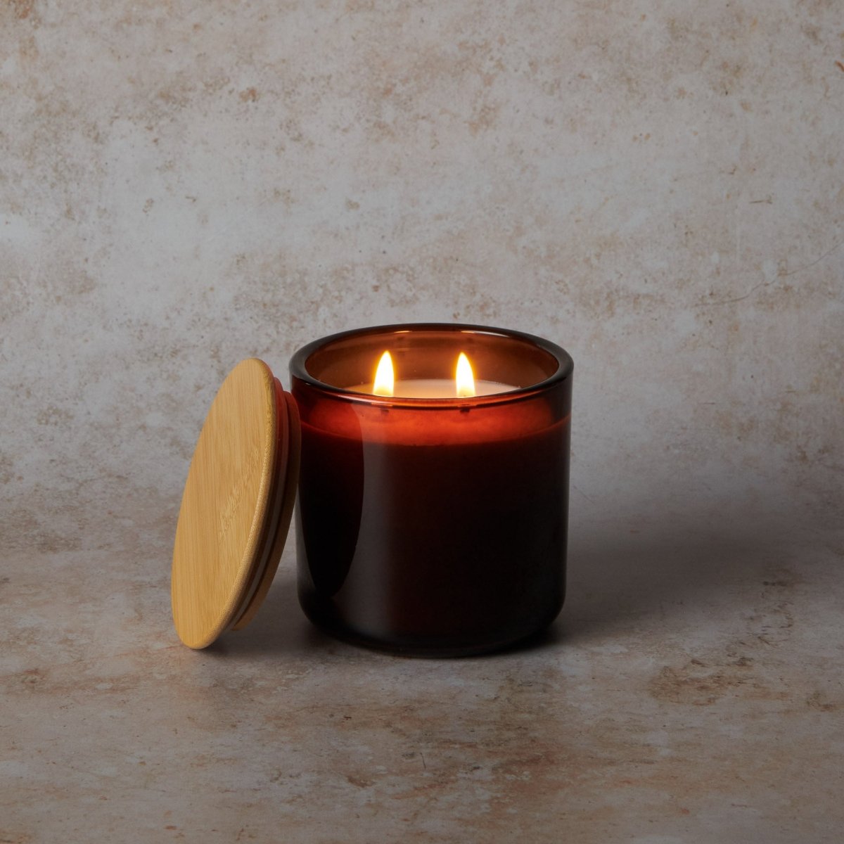 Lit candle in a dark glass jar with a wooden lid on a textured beige surface